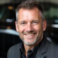 Porträt eines erfahrenen Ingenieurs, grinsend mit einem Schraubenschlüssel vor dem Hintergrund eines luxuriösen Sportwagens.